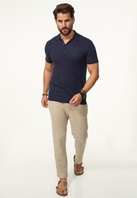 Marineblaue Polohemden mit kurzen Ärmeln, strukturiertem Stoff und einer Knopfleiste mit zwei Knöpfen; kombiniert mit hellbeigen strukturierten Hosen und braunen Sandalen.