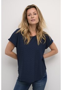 Blouse bleu marine à manches courtes avec un motif œillet complexe et un col rond ; associée à un jean en denim. Coupe droite, tissu léger.