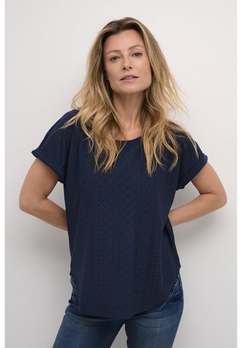Blouse bleu marine à manches courtes avec un motif œillet complexe et un col rond ; associée à un jean en denim. Coupe droite, tissu léger.