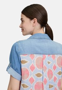 Koszula dżinsowa z krótkimi, podwiniętymi rękawami, niebieski przód i wzorzysty jasnoniebieski tył z różowymi i brązowymi kształtami w diamenty.