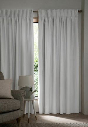 Tende grigio chiaro parzialmente aperte su una finestra, con una poltrona beige, un piccolo tavolino rotondo, una lampada e una piccola pianta in una stanza dai toni neutri.