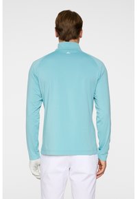 Ljusblå långärmad skjorta med höga krage och raglanärmar, tillverkad av ett mjukt tyg. Har en liten logotyp på baksidan.