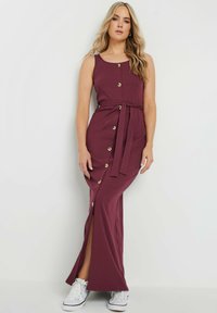 Robe maxi bordeaux avec une texture côtelée, dotée de boutons dorés, d'une taille ceinturée et d'une fente sur le côté, associée à des baskets blanches.