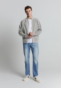 Jonge man staat met gevouwen handen, draagt een lichtgrijze zip-up jas, een wit T-shirt, lichtblauwe spijkerbroek en witte sneakers met marineblauwe accenten.