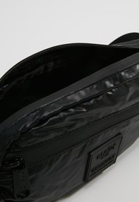 Czarna torebka crossbody wykonana z błyszczącego, teksturowanego materiału z zamkiem błyskawicznym i naszywką z logo. Wnętrze ma linię w paski.