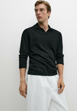 Mann trägt ein schwarzes Langarmhemd mit Kragen und weiße Hose, steht vor einem einfarbigen hellen Hintergrund und schaut zur Seite.