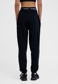 Zwarte joggingbroek van zacht materiaal met een elastische tailleband en witte branding. Tapere pijpen met boorden en zichtbare zijzakken.