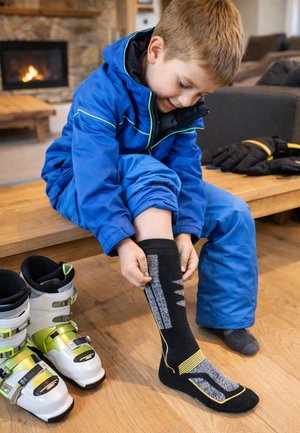 Kind in blauer Outdoor-Bekleidung sitzt auf einer Holzbank und zieht einen schwarzen Socke mit grauen und gelben Mustern an. Skischuhe sind in der Nähe.