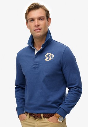 Blue long-sleeve polo shirt with a high collar, featuring a beige logo on the left chest and a white placket. Worn with a brown braided belt.