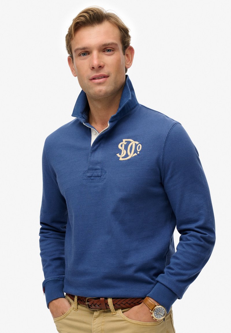 Blue long-sleeve polo shirt with a high collar, featuring a beige logo on the left chest and a white placket. Worn with a brown braided belt.