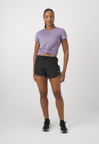 Purple short-sleeve sports top with knotted front, paired with black shorts featuring an elastic waistband and small logo detail. Black athletic shoes.