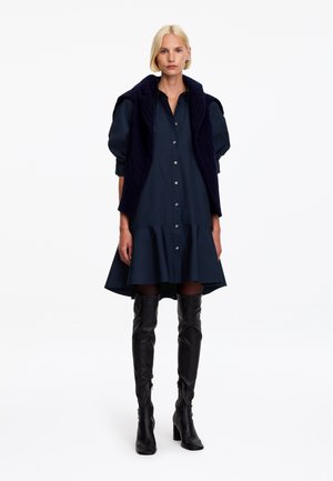 Femme blonde portant une robe bleu marine boutonnée avec des manches ballons, un gilet sombre sans manches et des bottes noires montantes cuisses, debout sur un fond blanc.