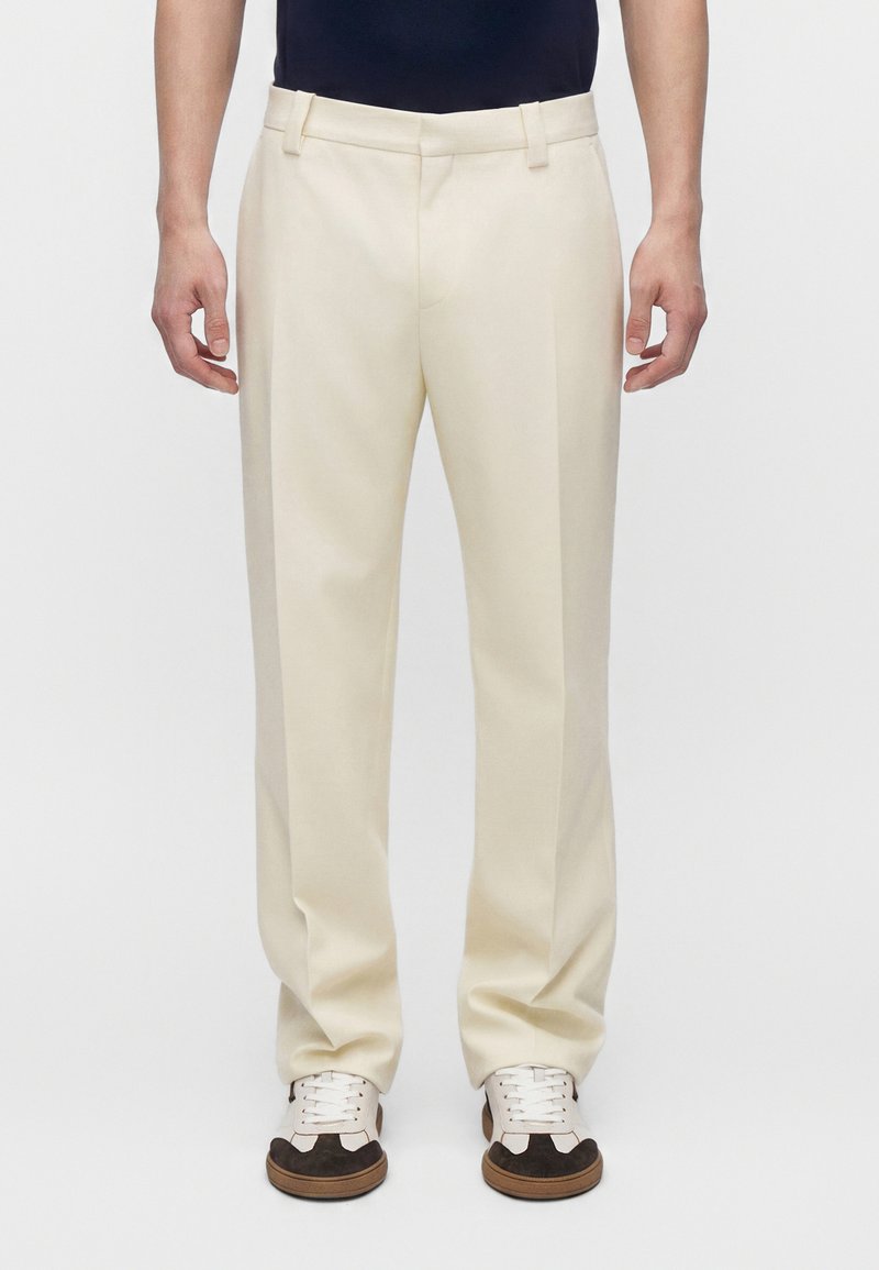 Homme portant un pantalon couleur crème et des baskets blanches et marron, debout devant un fond uni clair.