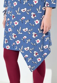 Blaue Tunika mit einem floralen Muster in Weiß und Rot, mit einem diagonalen Saum. Getragen über bordeauxfarbenen Leggings aus weichem Stoff.