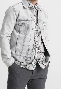 Homme portant une veste en denim gris clair par-dessus une chemise blanche avec un motif de cactus noir et rouge et un pantalon gris foncé, les mains dans les poches.