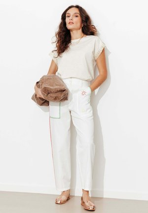 Femme aux longs cheveux bouclés portant un haut crème et un pantalon blanc avec une broderie colorée, tenant une veste marron pliée contre un mur blanc.