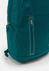 Teal backpack with a textured fabric, featuring a side zippered pocket and a reflective strip. Includes adjustable straps and a bottom loop.