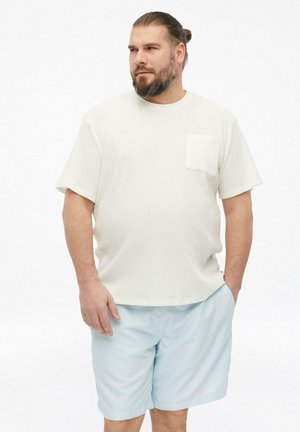Man met baard en naar achter gebonden haar draagt een wit T-shirt met korte mouwen en lichtblauwe korte broek, staat met één hand in de zak en kijkt opzij.