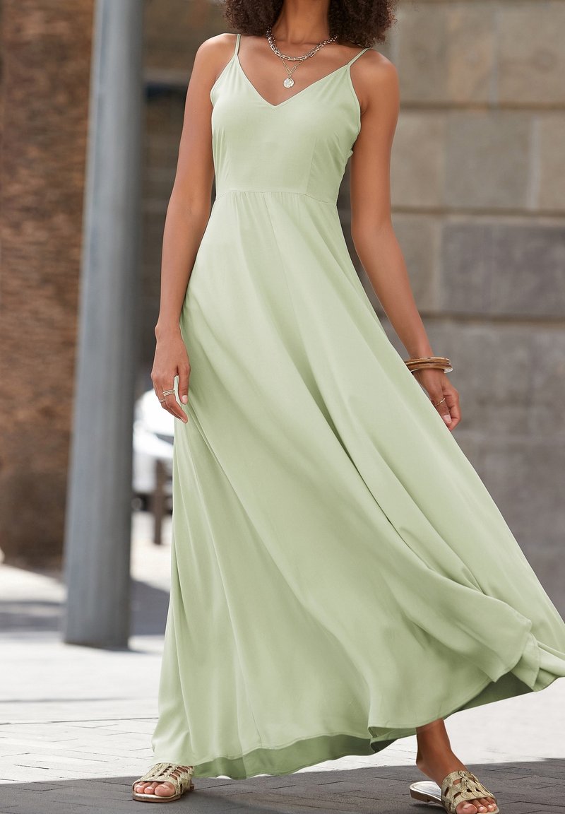 Femme portant une robe maxi sans manches vert clair avec une jupe fluide, des colliers superposés, des bracelets et des sandales tressées, debout en extérieur.