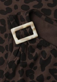 Tissu marron avec imprimé léopard noir et une boucle de ceinture rectangulaire beige marbrée attachée à une sangle en tissu.