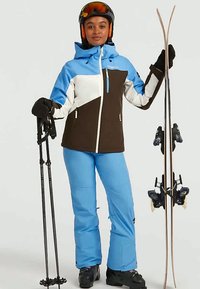 Ski outfit featuring a blue and white jacket with brown accents, paired with light blue pants. Ski poles in one hand, skis in the other.