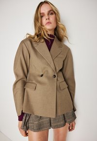 Blazer castanho de dupla face com lapelas em bico, dois bolsos frontais e botões pretos. Usado sobre um suéter de gola alta em ponto canelado na cor borgonha e calções de xadrez.