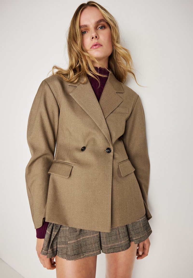 Blazer castanho de dupla face com lapelas em bico, dois bolsos frontais e botões pretos. Usado sobre um suéter de gola alta em ponto canelado na cor borgonha e calções de xadrez.