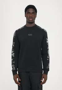 Sweatshirt preto com mangas longas, apresentando designs gráficos em branco nos ombros e um patch de logotipo texturizado na parte da frente.