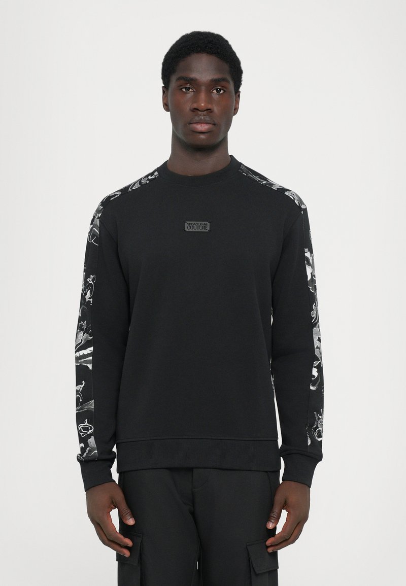Sweatshirt preto com mangas longas, apresentando designs gráficos em branco nos ombros e um patch de logotipo texturizado na parte da frente.