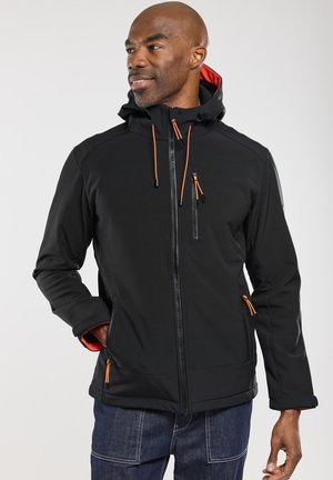 Homme portant une veste noire à capuche avec des tirettes de zip orange et un jean bleu, debout avec une main dans la poche, regardant vers la gauche.