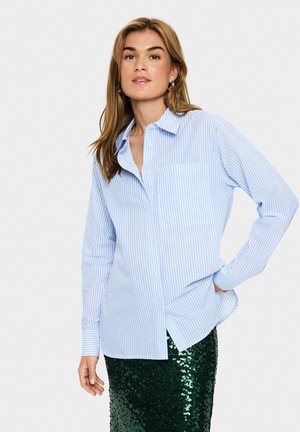 Light blue and white striped button-up shirt with a single chest pocket, long sleeves, and a relaxed fit, worn over a green sequin skirt.