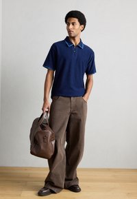 Άνδρας που φοράει μπλε σκούρο κοντομάνικο πόλο με μπλε λεπτομέρειες, καφέ παντελόνι φαρδύ πόδι, σκούρα παπούτσια, κρατώντας μία μεγάλη καφέ δερμάτινη τσάντα, στέκεται σε ξύλινο πάτωμα.