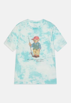 Jasnoniebieski T-shirt typu tie-dye z misiem w sprzęcie wędkarskim, czerwoną czapką, trzymającym wędkę, z napisem „Polo Bear by Ralph Lauren” poniżej.