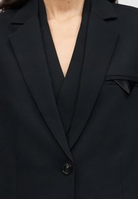 Γυναίκα που φοράει μαύρο σακάκι με ένα κουμπί και ένα ταιριαστό διπλωμένο μαντηλάκι στην τσέπη αριστερά.