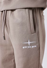 Light brown, soft fabric joggers featuring an elastic waistband, drawstring closure, and prominent white embroidered logo on the thigh.