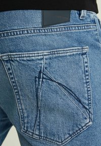 Šviesiai mėlyni džinsai su išskirtiniu kišenės dizainu ir odiniu užsegimu prie juosmens. Tekstūruotas audinys su matomais siūlėmis.