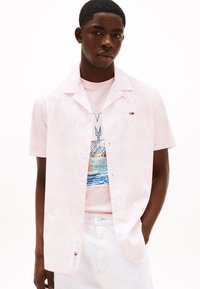 Camicia a maniche corte di colore rosa chiaro con un motivo sottile, realizzata in un tessuto morbido. Presenta una chiusura frontale con bottoni e un piccolo logo sul petto.