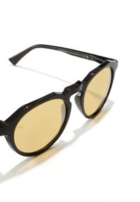 Musta päikeseprillid tekstuurse raamiga, ümmarguste kollaste läätsedega ja nurkadega disainielementidega. Raami materjalis on peen sädelus.
