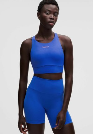 Mujer atlética de pie, vistiendo un sujetador deportivo azul a juego y pantalones cortos para ciclismo de cintura alta sobre un fondo gris liso.