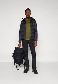 Fekete szigetelt dzseki kapucnival, zöld középréteg, fekete nadrág és fekete hátizsák. Kis logóval és masszív csizmákkal rendelkezik. Simára tapintó anyag.