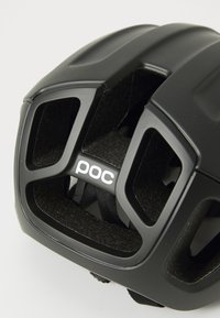 Czarny kask rowerowy z licznymi otworami wentylacyjnymi, teksturowanym wnętrzem, gładką powierzchnią zewnętrzną oraz białym logo marki "poc" z boku.