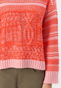 Knit sweater featuring a mix of coral and pink tones, textured patterns, and ribbed hem, paired with olive green pants.