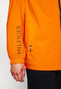 Manche de veste orange vif avec poignet élastique et texte noir "HILFIGER" imprimé verticalement, ouverture de poche, et une partie d’une main ainsi que d’un pantalon noir.