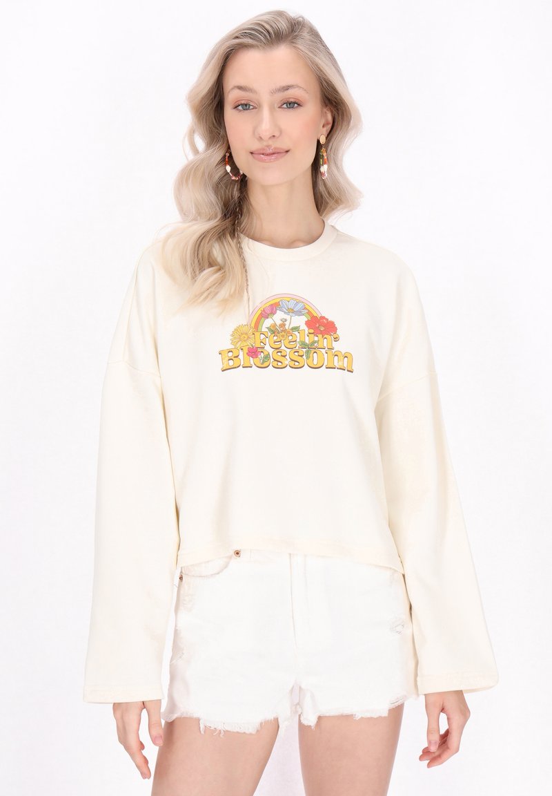 Sweatshirt cropped a maniche lunghe color crema con una grafica floreale e arcobaleno colorata con il testo "Blossom". Abbinato a pantaloni short bianchi strappati.