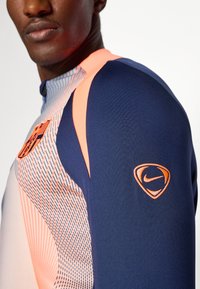 Haut de sport Nike en bleu et pêche. Présente un design élégant, des motifs texturés et un logo emblématique sur la manche. Lignes épurées, style ajusté.