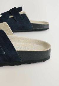 Tamno plave suede sandale s mekim bež uložkom, podesivim remenom i hrapavom crnom gumenom potplatom; dizajn s otvorenim prstima.