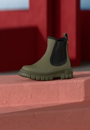 Groene rubberen enkellaars met een zwart elastische zijpaneel, een getextureerde zool en een trekband. Gemaakt tegen een koraalkleurige trap achtergrond.
