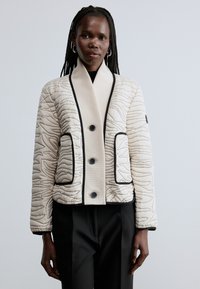 Sieviete valkā krēmkrāsas izšūtu jaku ar melnu apmali un pogām, kombinezonā ar melnām biksēm, stāvot pret vienkāršu gaiši pelēku fonu.