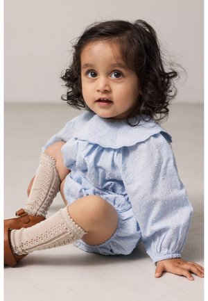 Νήπιο με καστανά σγουρά μαλλιά, φοράει ανοιχτό μπλε ρούχο, κρεμ πλεκτές κάλτσες μέχρι το γόνατο και καφέ παπούτσια, καθισμένο σε ανοιχτό δάπεδο.