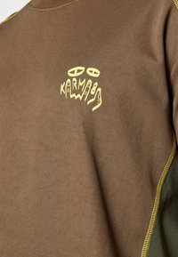 Sweatshirt em mistura de algodão castanho com costura amarela e um logótipo gráfico que diz "KARMA89" no peito.